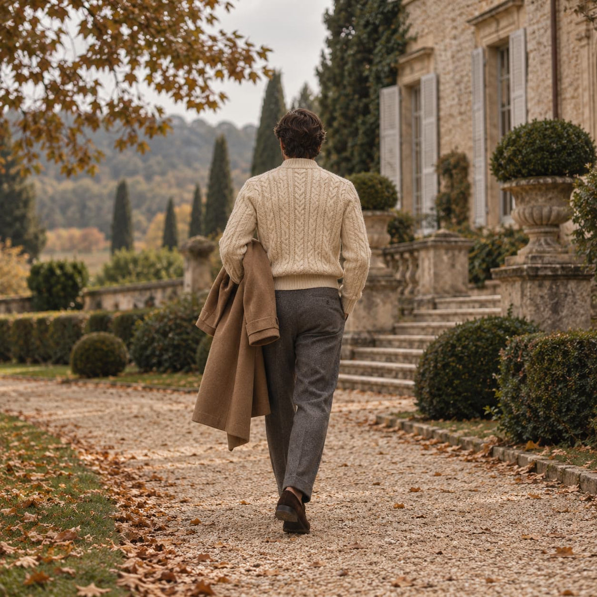 Homme de dos dans une allée de jardin, pull crème, manteau camel sur le bras et pantalon gris